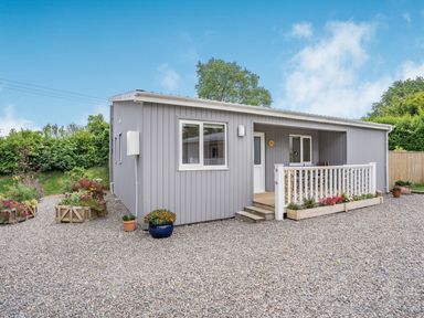 A small grey lodge with a front porch, flower boxes, and gravel driveway at Caldey Lodge  Bier Wood Lodges in Manorbier