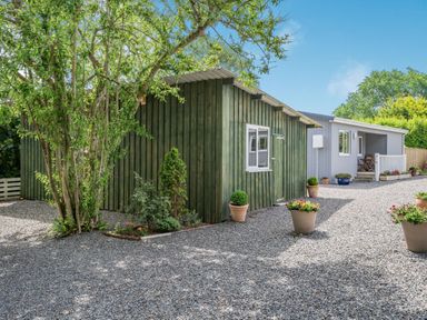 An outdoor area with a gravel ground two wooden buildings a tree and several potted plants at Caldey Lodge Bier Wood Lodges in Manorbier