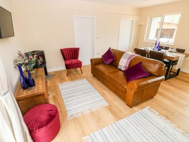 A living room with a brown leather sofa purple cushions red armchair wood stove dining table and chairs at Caldey Lodge Bier Wood Lodges Manorbier