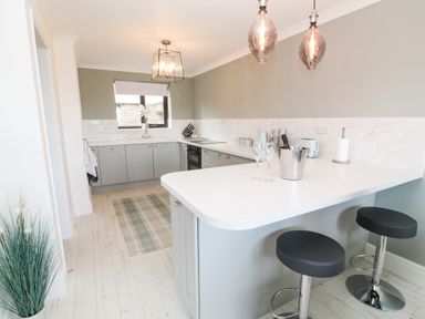 A kitchen with grey cabinets a white countertop bar with two stools and a window at 12 Claremont Falls Polperro