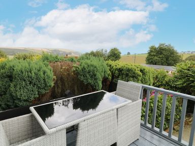 An outdoor seating area with a glass table and two wicker chairs on a deck with bushes and countryside in the background at 12 Claremont Falls in Polperro