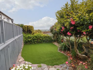 A small garden with a green lawn bordered by flowering trees and shrubs at 12 Claremont Falls in Polperro