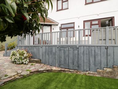A garden with artificial grass next to a stone pathway and a wooden deck attached to a white house at 12 Claremont Falls in Polperro