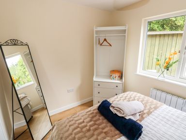 A bedroom with a bed folded towels a mirror a white wardrobe with hangers and a window with flowers on the sill at Ramsey Lodge Bier Wood Lodges in Manorbier