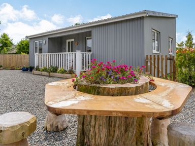An outdoor area with a grey cabin a wooden table with flowers in the center and wooden stools at Ramsey Lodge Bier Wood Lodges in Manorbier