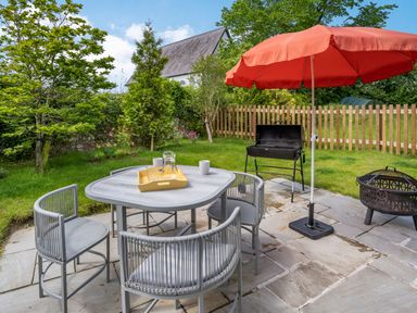 A patio with a table and four chairs a red umbrella a barbecue grill and a fire pit with a grassy fenced garden in background at Ramsey Lodge Bier Wood Lodges in Manorbier