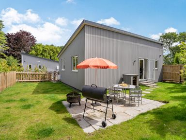 A backyard with a patio table and chairs an orange umbrella a barbecue grill and a fire pit at Ramsey Lodge Bier Wood Lodges in Manorbier