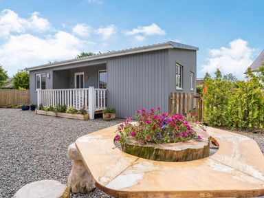 A grey lodge with white porch railing surrounded by gravel and a wooden bench with flowers in the center at Ramsey Lodge Bier Wood Lodges in Manorbier