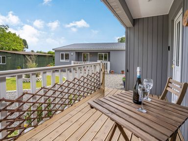 A wooden deck with a table bottle and two glasses overlooking a gravel yard and a gray house at Ramsey Lodge Bier Wood Lodges in Manorbier