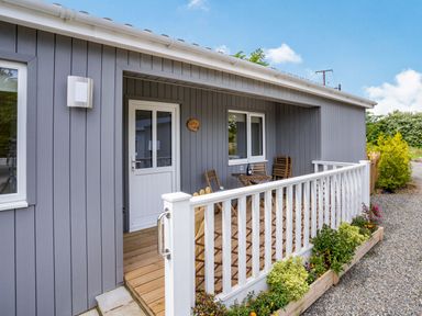 A porch with wooden chairs and table outside a grey house with white door and window at Ramsey Lodge Bier Wood Lodges in Manorbier