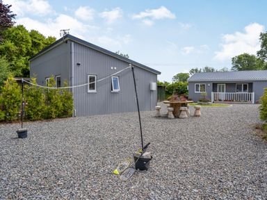A yard with a badminton net and rackets between two gray buildings with a stone table and stools at Ramsey Lodge Bier Wood Lodges in Manorbier