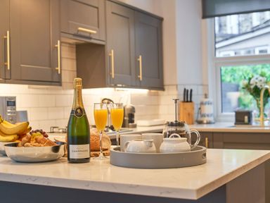 A kitchen countertop with a bottle of champagne two glasses of orange juice a bowl of fruit and pastries and a tray with cups at Lower Sheriff's Place in Windermere