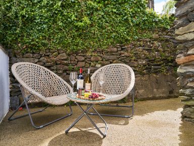Two wicker chairs with a small round glass table holding two wine bottles grapes and jars against a stone wall with ivy at Lower Sheriff's Place in Windermere