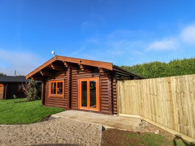 A wooden log cabin with glass doors and a wooden fence in a grassy area at Oak Lodge in Greetham Rutland