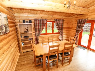 A dining area with a wooden table and six chairs a wooden bookshelf with decor and a window with plaid curtains at Oak Lodge in Greetham Rutland