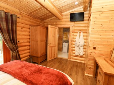 A bedroom with a red blanket on the bed wooden furniture robes hanging on the wall and a TV mounted at Oak Lodge in Greetham Rutland