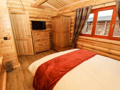 A bedroom with wooden walls a bed with a red runner a wooden chest of drawers a wardrobe and a window with curtains at Oak Lodge in Greetham Rutland