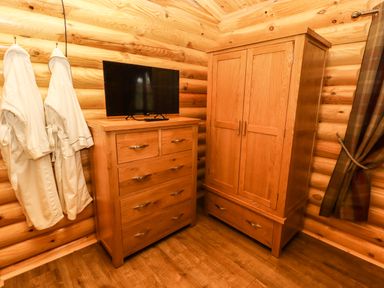 A corner of a room with wooden chest of drawers with a television on top a wooden wardrobe two white robes hanging on the wall and a plaid curtain on a wooden log wall at Oak Lodge in Greetham Rutland