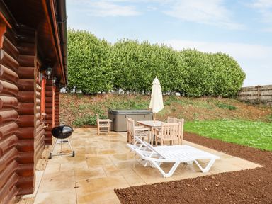 An outdoor patio with wooden dining table and chairs a barbecue grill and a white loungers near a wooden cabin at Oak Lodge Greetham Rutland