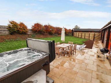 An outdoor patio with a hot tub wooden dining table with chairs two loungers barbecue grill and a lawn with trees at Oak Lodge in Greetham Rutland