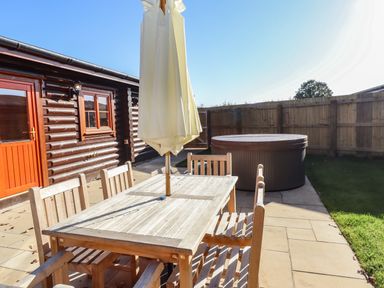An outdoor patio with a wooden table and chairs and a closed umbrella next to a hot tub and a wooden fence at Holly Lodge in Greetham Rutland