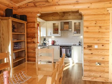 A wooden dining table with chairs next to a bookshelf and an open kitchen with white cabinets at Holly Lodge in Greetham Rutland
