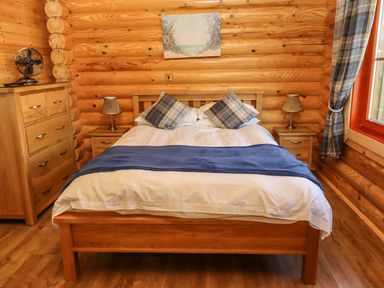 A bedroom with a wooden bed a blue throw and plaid cushions two bedside tables with lamps a chest of drawers and a window with plaid curtains at Holly Lodge in Greetham Rutland