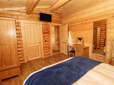 A bedroom with wooden walls and ceiling beams a bed with blue blanket a wooden wardrobe a vanity table with mirror and stool a full-length wall mirror and a wall-mounted television at Holly Lodge in Greetham Rutland