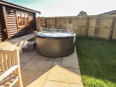 A hot tub on a paved patio beside a wooden house with a fenced lawn at Holly Lodge in Greetham Rutland