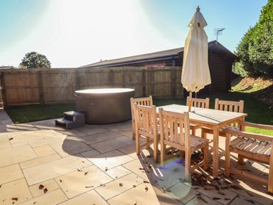 An outdoor patio with wooden table, chairs, a closed umbrella, and a hot tub with steps at Holly Lodge in Greetham Rutland
