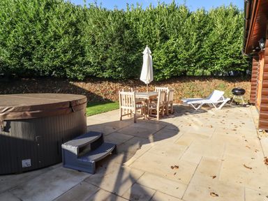 An outdoor patio with a hot tub a table with six chairs and an umbrella a sun lounger and a barbecue grill at Holly Lodge in Greetham Rutland