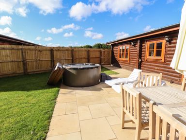 An outdoor patio area with wooden table and chairs white lounge chairs hot tub and wooden fence next to a log cabin at Pine Lodge in Greetham Rutland
