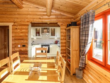 A dining area with wooden table and chairs next to a kitchen with white cabinets and a wooden bookshelf by a window with plaid curtains at Pine Lodge in Greetham Rutland