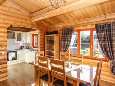 A dining area with a wooden table and chairs next to windows with plaid curtains and a kitchen in the background at Pine Lodge in Greetham Rutland