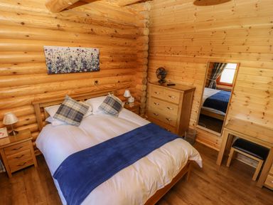A bedroom with a double bed with pillows and a blue blanket a wooden dresser a mirror a small table and wooden walls at Pine Lodge in Greetham Rutland