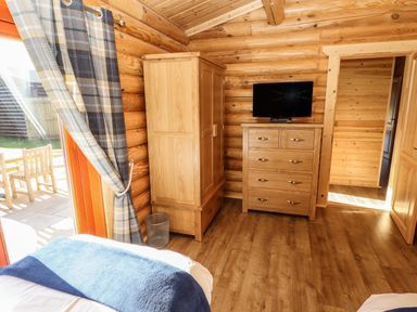 A bedroom with wooden walls and floor a wardrobe a chest of drawers with a television and a window with plaid curtains at Pine Lodge in Greetham Rutland