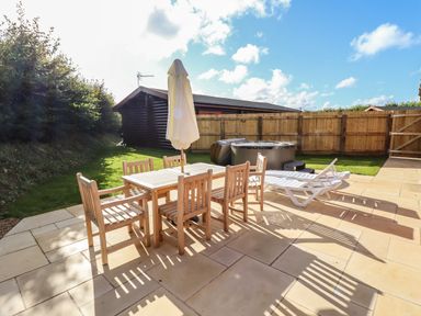An outdoor patio area with a wooden table and six chairs a closed umbrella a white lounge chair a hot tub and a wooden fence at Pine Lodge in Greetham Rutland