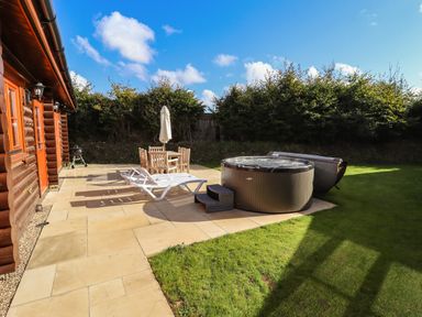 An outdoor patio with lounge chairs a table with chairs and umbrella and a hot tub next to a lawn at Pine Lodge in Greetham Rutland