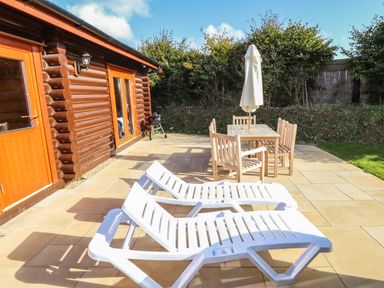 An outdoor patio area with two white lounge chairs a wooden dining table with four chairs and a closed umbrella next to a log cabin at Pine Lodge in Greetham Rutland
