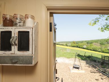 A wall-mounted cabinet with glass jars inside a room with an open door overlooking a green field and a tripod barbecue at The Shepherds Hut at Marley in Marley near Barham Kent