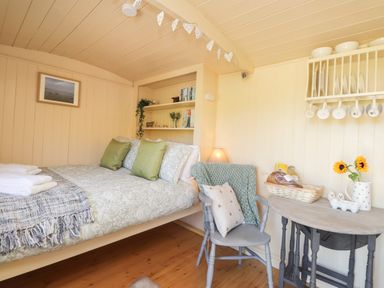 A small bedroom with a bed with green pillows a chair a small table with a basket and sunflowers and shelves with dishes and books at The Shepherds Hut at Marley