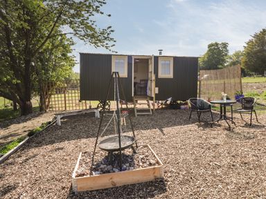 An outdoor area with a small black hut on wheels a fire pit with a grill a bench and a round table with two chairs at The Shepherds Hut at Marley Marley near Barham Kent
