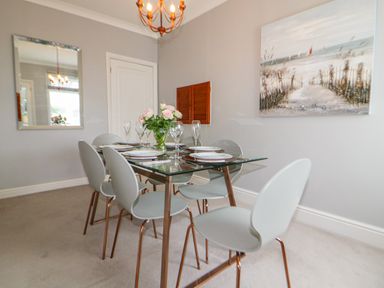 A dining room with a glass table set with plates and glasses six chairs a mirror and a beach painting on the wall at Robin Hill in Duporth near St Austell