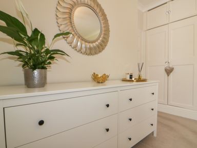 A white dresser with decorative items a plant and a round mirror above in a room at Robin Hill in Duporth near St Austell