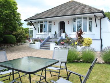 A house with stairs leading to the entrance and a glass table with chairs on the lawn at Robin Hill in Duporth near St Austell