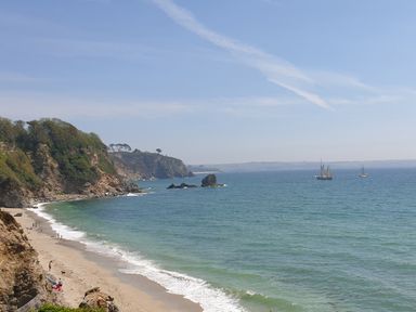 A sandy beach with people walking near cliffs and boats sailing on the sea at Robin Hill in Duporth near St Austell