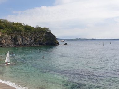 A beach with people swimming and sailing near a rocky cliff and calm sea at Robin Hill in Duporth near St Austell