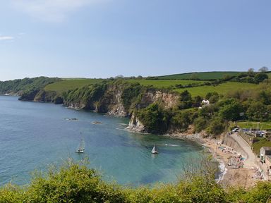 A coastal scene with cliffs green fields sailboats and a small beach with people at Robin Hill in Duporth near St Austell