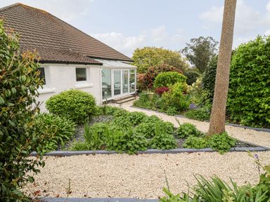 A garden with a gravel path and various green bushes and plants next to a white house with a brown roof at Robin Hill in Duporth near St Austell