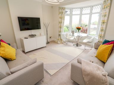 A living room with two beige sofas a round glass table with chairs a wall-mounted TV and bay windows with floral curtains at Robin Hill in Duporth near St Austell